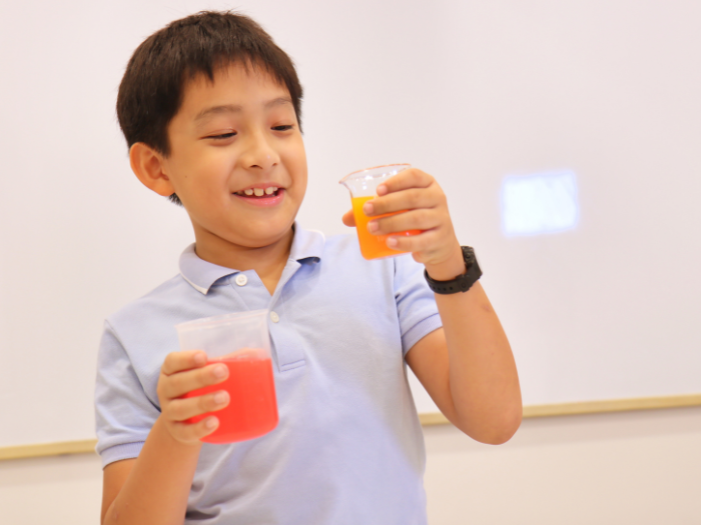 Boy in science class, holding two beakers with bright pink and orange content